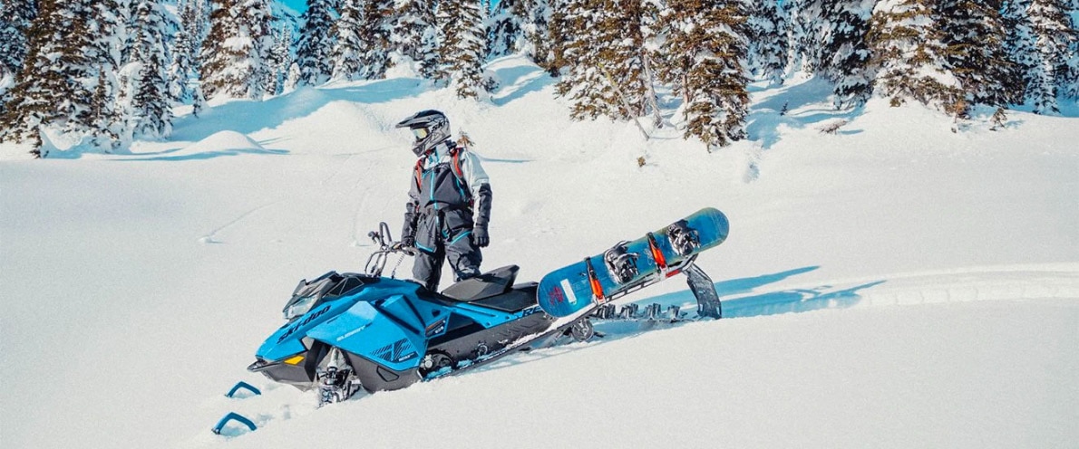 Muž vedle svého sněžného skútru Ski-Doo Summit se snowboardem