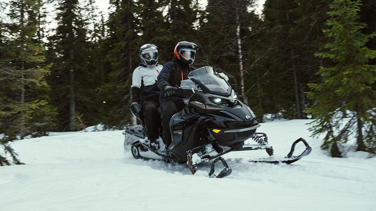 Two people riding a Lynx Adventure snowmobile in a snowy forest