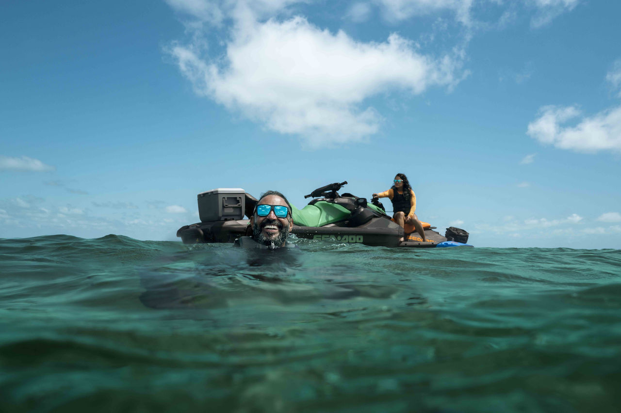A man wearing polarized sunglasses in the water with two Sea-Doo personal watercraft behind him, with a woman sitting on one of it