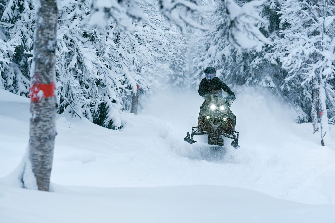 Райдер выполняет прыжок на снегоходе Lynx Xterrain RE 2027 в заснеженном лесу