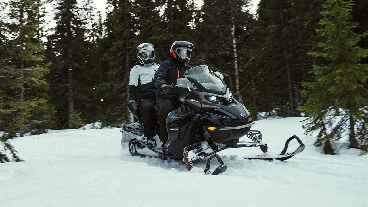 Двое едут на снегоходе Lynx Adventure по заснеженному лесу