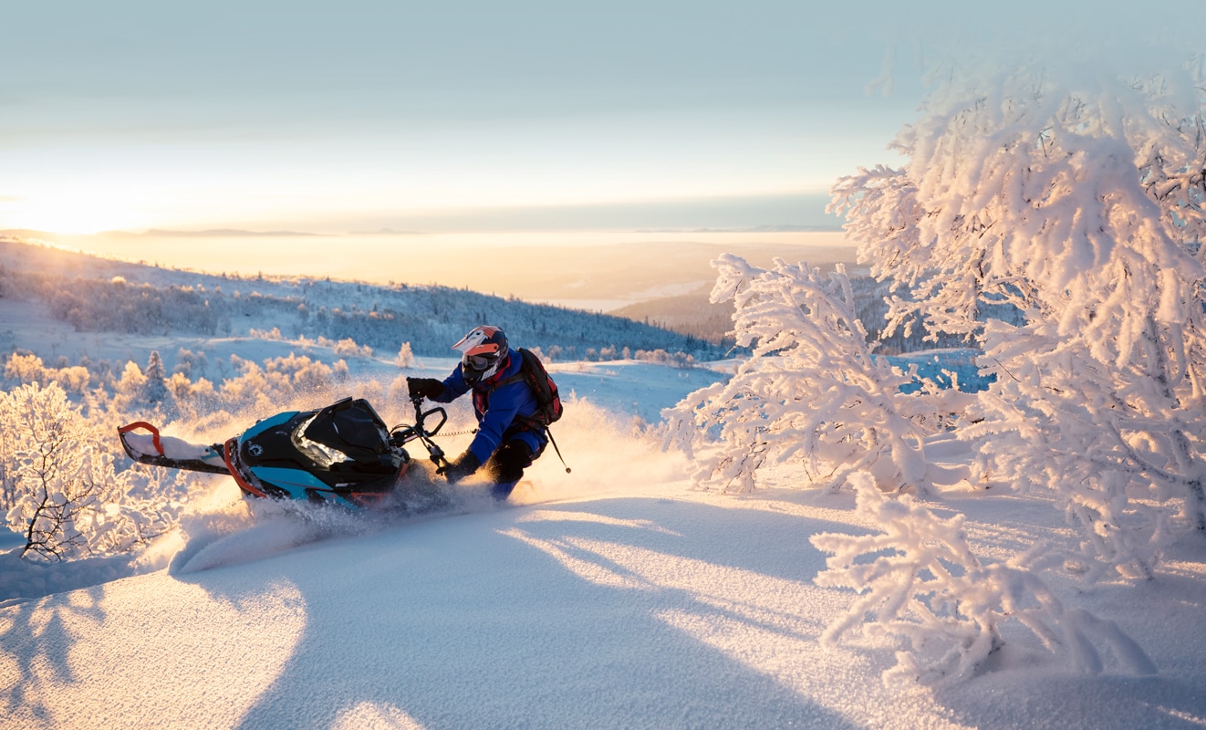 Muž prudce zatáčející svůj sněžný skútr Lynx BoonDocker 3900 na zasněženém kopci při západu slunce