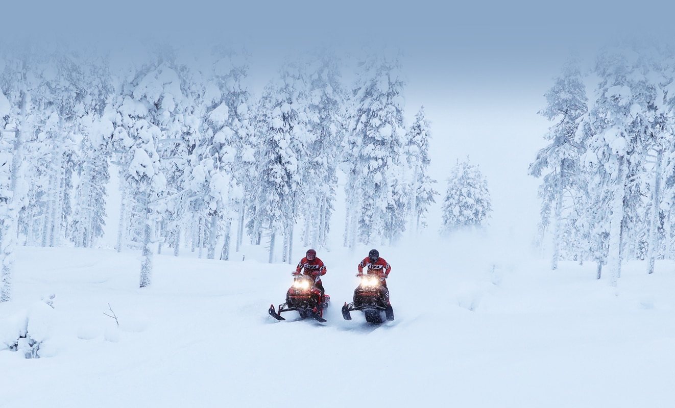 Two men drive on a snowy road through the forest with their Lynx Rave Re Snowmobile Model