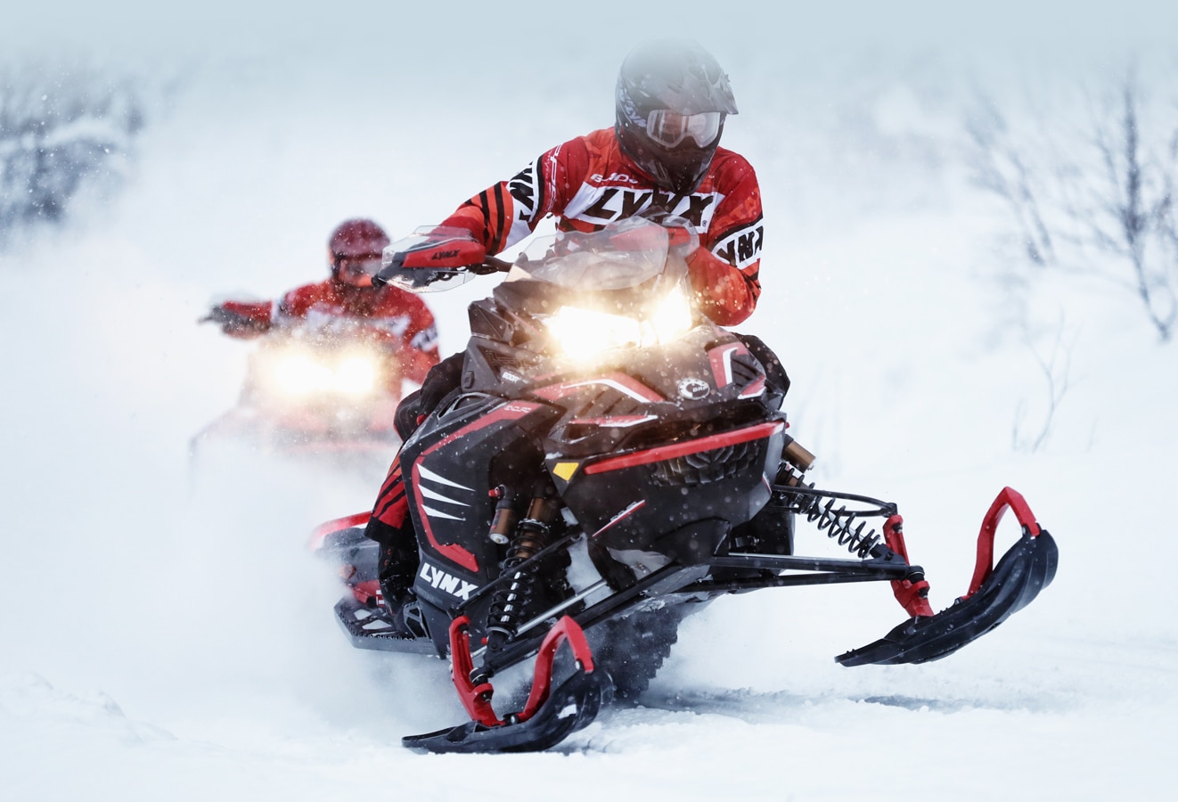 A man is driving the red Lynx Rave Re Snowmobile Model and a man is following him. 
