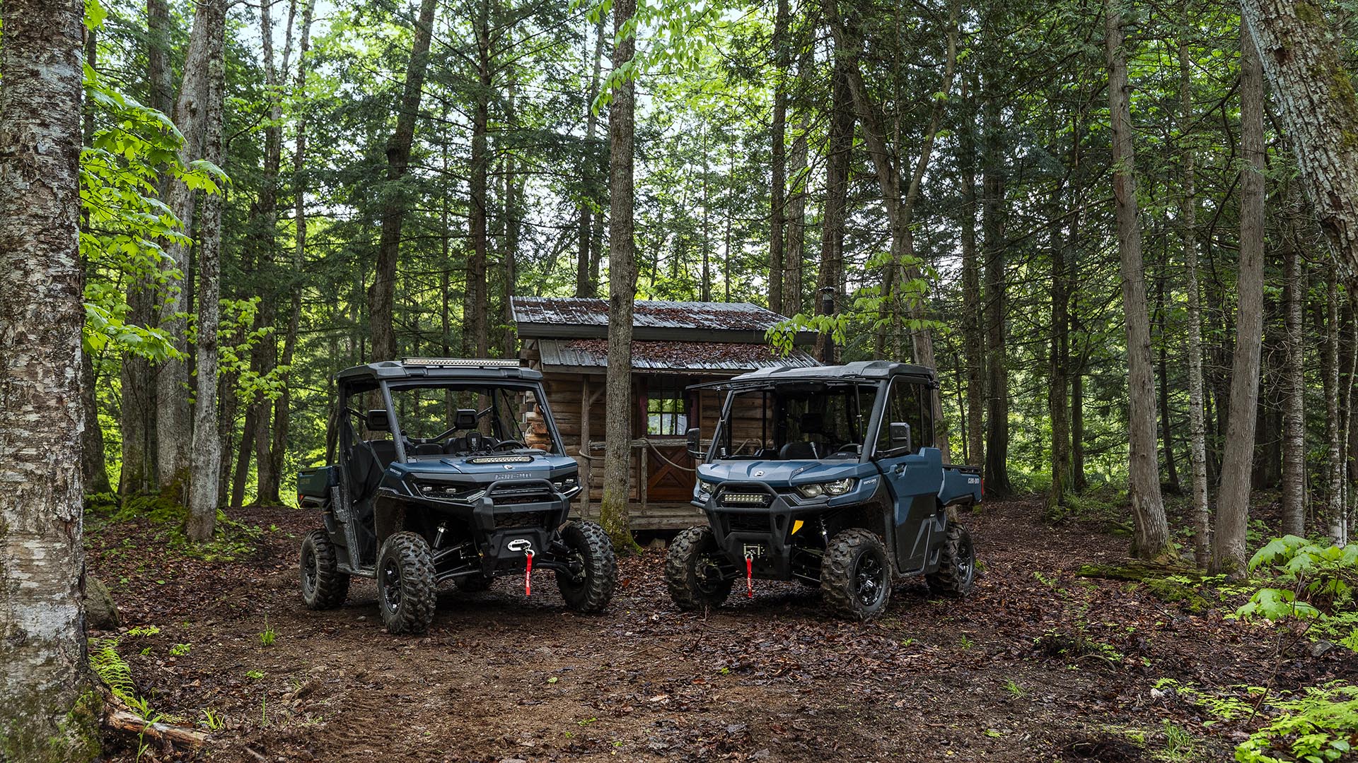 Two 2026 Can-Am Traxter parked in a forest near a cabin