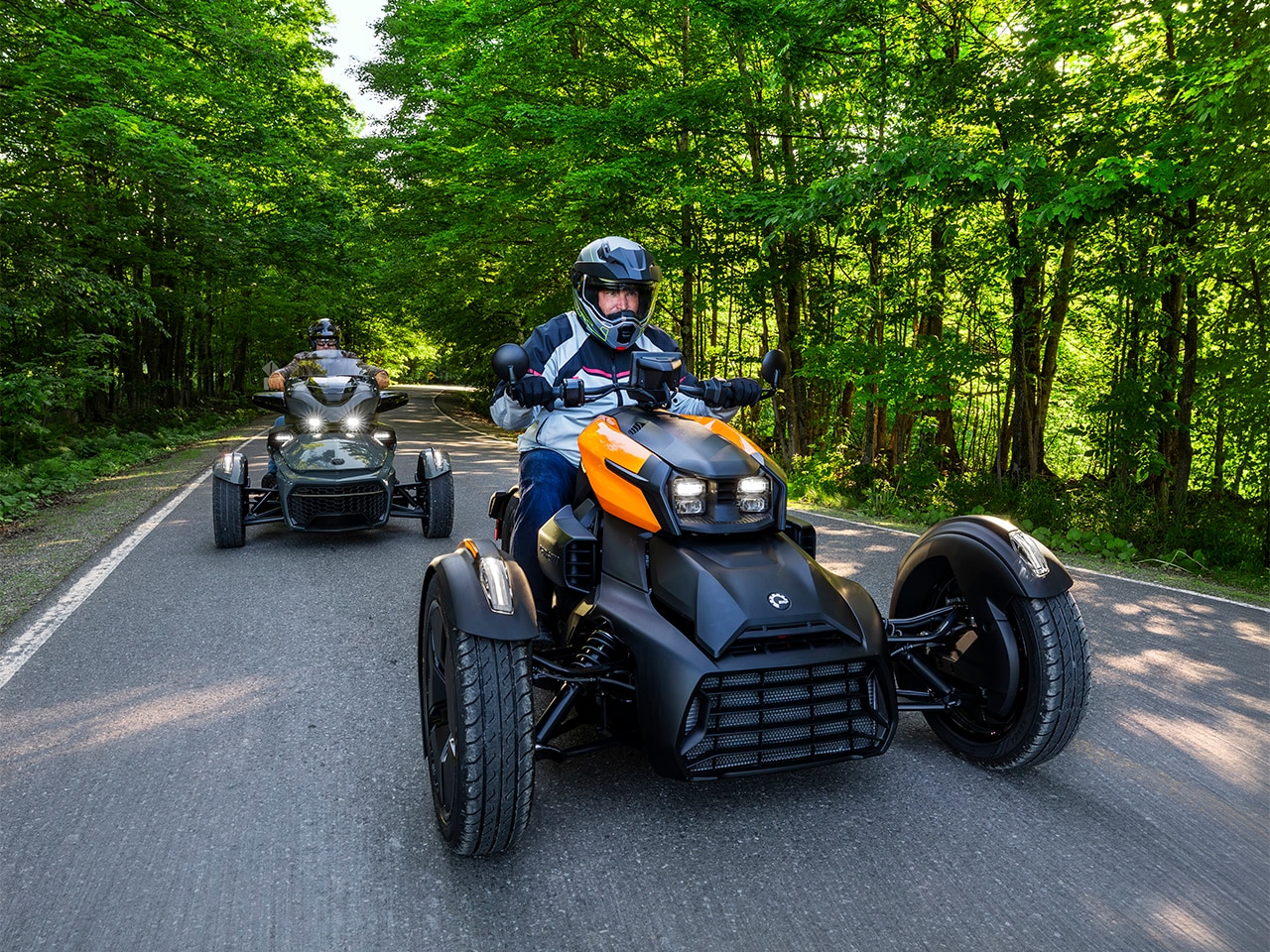 A 2027 Can-Am Ryker followed by a 2027 Can-Am Spyder on a scenic tree-lined road