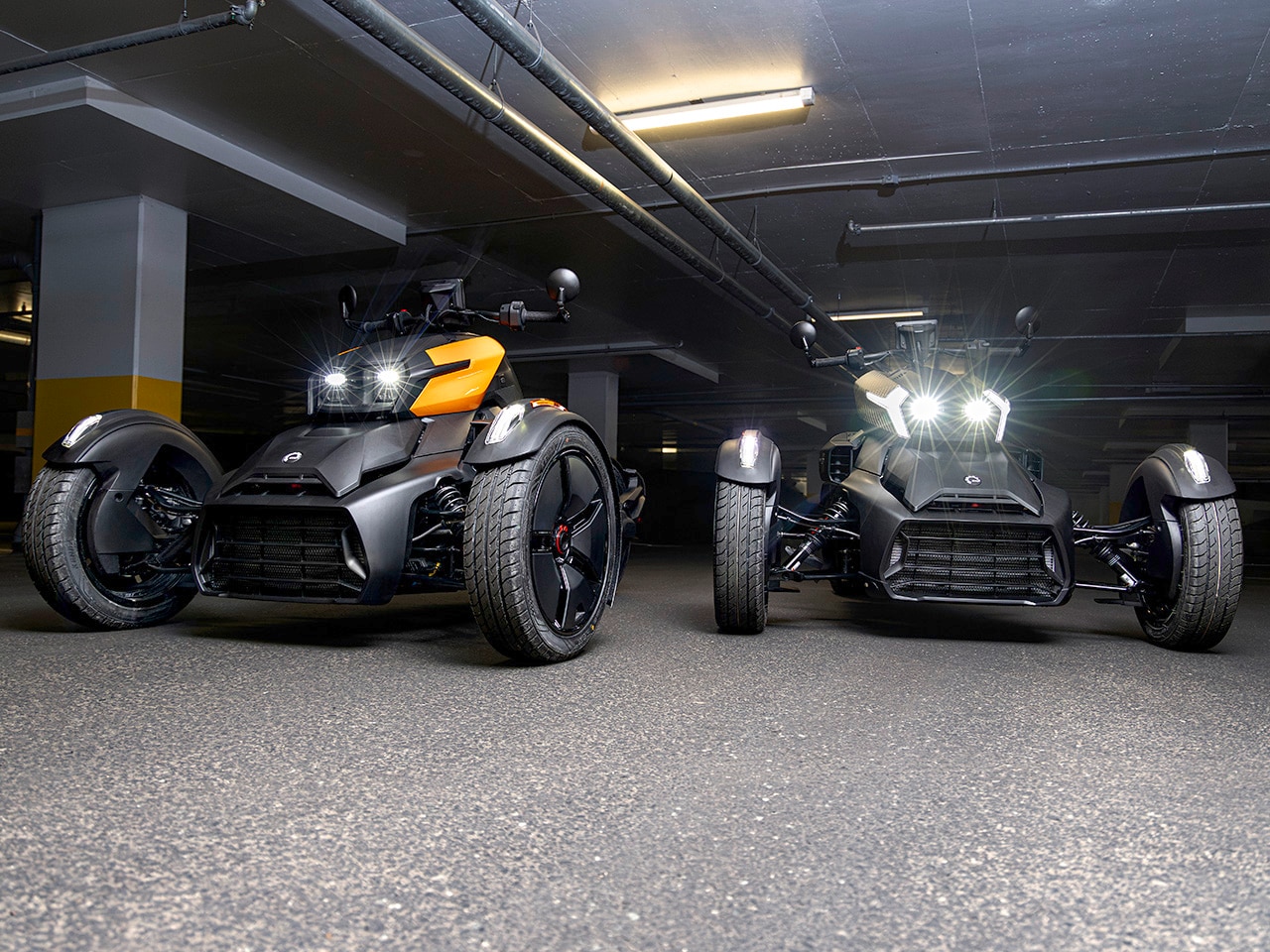 Two 2027 Can-Am Ryker 3-wheel vehicles with their headlights on in a parking garage