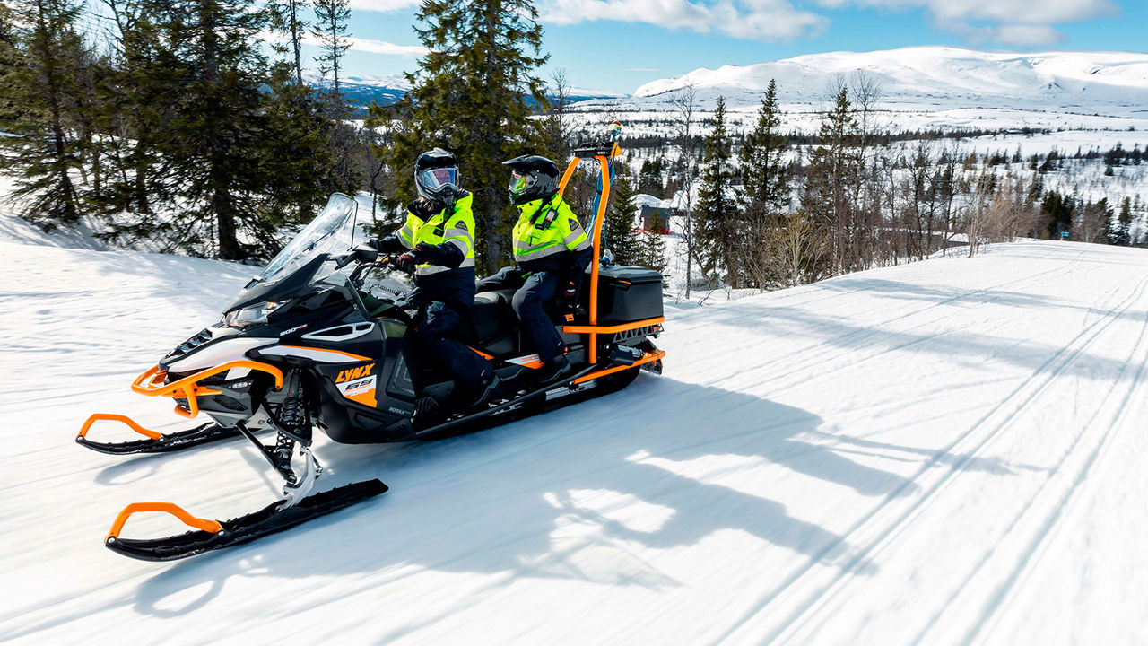 Workers riding Lynx 69 Ranger Alpine snowmobile uphill in ski slope