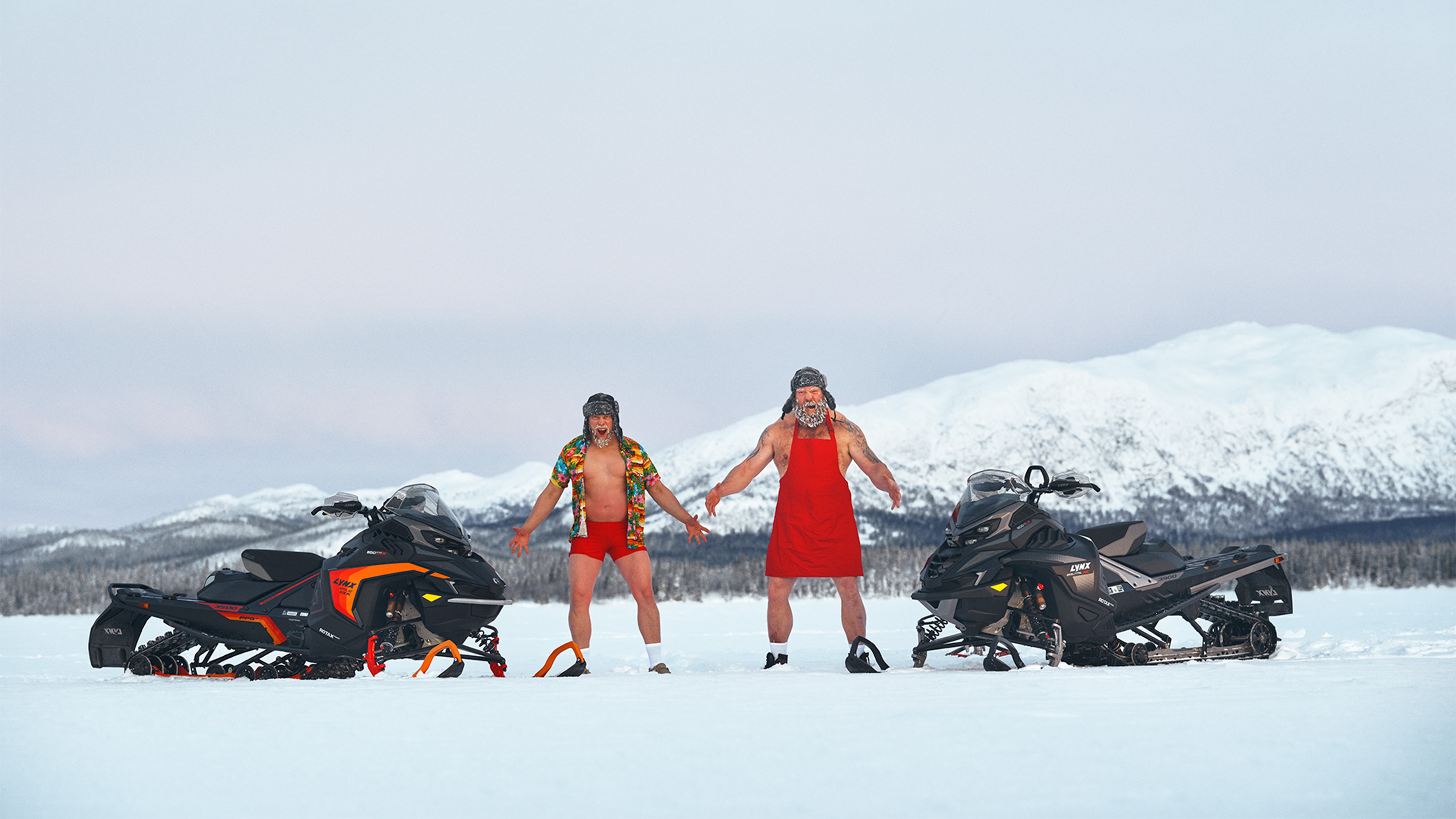 Two men roaring in front of Lynx Rave GLS and Lynx Brutal RE 2025 snowmobiles on lake ice