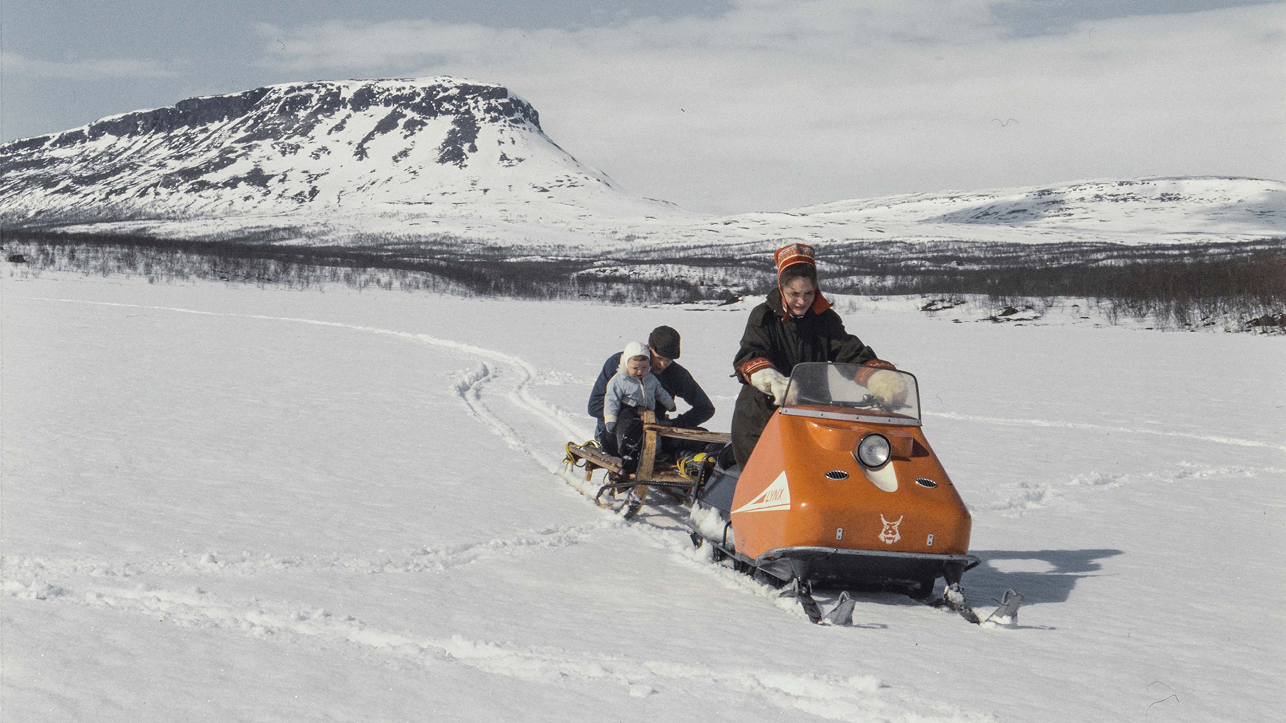 Žena ťahá muža a dieťa na saniach v Kilpisjärvi so snežným skútrom skútrom Lynx AS 50 1968